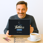 A smiling man in a dark sweater holds a navy Hattie’s Gifts ‘Happy Father’s Day’ hamper box on a light wood table, with a white cup of coffee and a sprig of flowers beside it, against a plain pale background